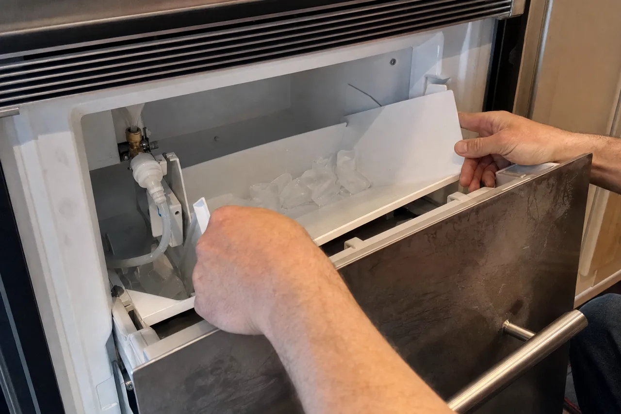 The open drawer of a built-in ice maker with the bin withdrawn and the water fittings visible behind it.