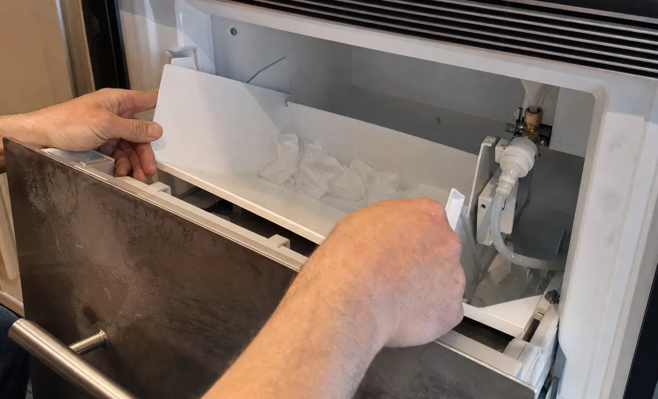 An ice maker drawer open at counter height, the bin half out and the water fitting in shadow behind.