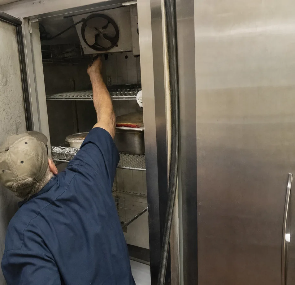 An arm reaching up to the evaporator fan housing at the top of an open refrigerated cabinet, corrosion staining the door frame beside it.