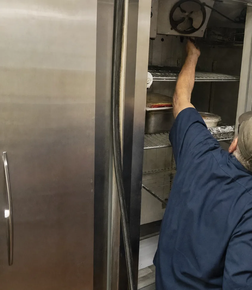 The open door of a stainless refrigerator held back while a hand works at the fan grille above the top shelf.