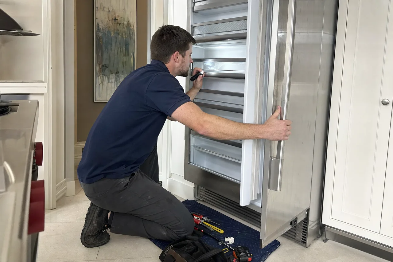 A technician kneeling at an open stainless built-in refrigerator, steadying the door with one hand and lighting the interior with a flashlight.