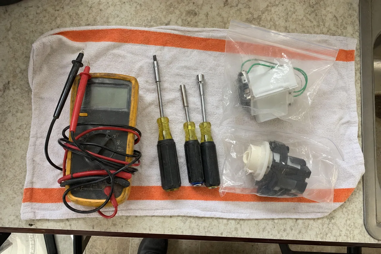 An overhead view of a technician’s tools on a kitchen towel: a multimeter, nut drivers and two replacement parts still in their bags.